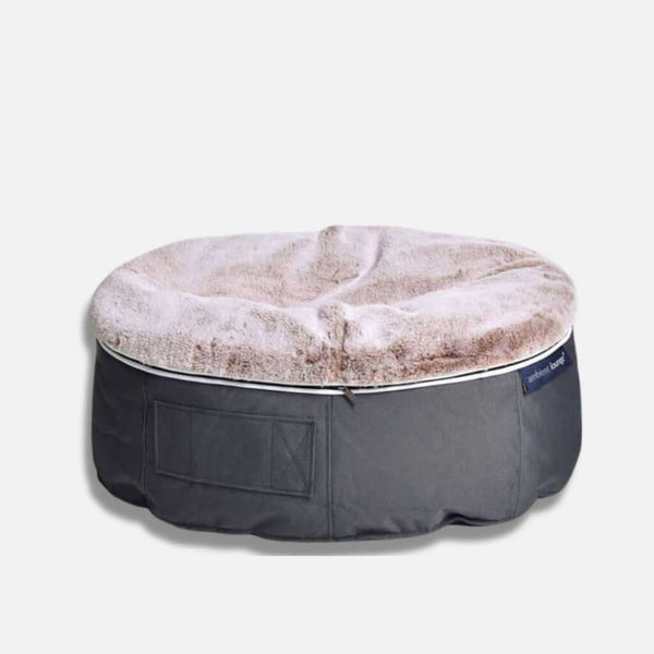 En rund Ambient Lounge hundeseng med en mjuk, beige, fluffig topp och en mörkgrå tygbotten, med en liten sidoficka och etikett på sidan, mot en ljus bakgrund.