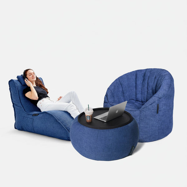 En kvinna kopplar av i en blå fåtölj med fötterna uppe, bredvid en matchande blå fåtölj och en rund ottoman, och skapar en mysig sittegrupp med sin laptop och drink, perfekt för att planera den ultimata hjemmekino mot en vanlig vit bakgrund.