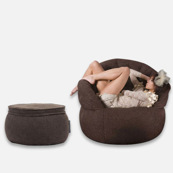 En lättklädd kvinna med hatt sitter uppkrupen i en stor, rund Butterfly Chaise-fåtölj. Bredvid henne står en matchande ottoman i memory foam, båda med ett mjukt, plyschigt utseende. Bakgrunden är helt vit.