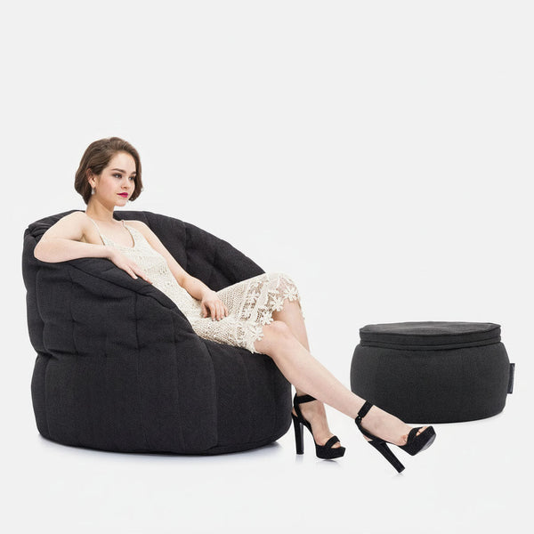 En kvinna i vit spetsklänning och svarta högklackade skor sitter med korsade ben på en stor, mörk Memory Foam Butterfly Sofa. En matchande rund ottoman är placerad bredvid henne, allt mot en vanlig vit bakgrund.