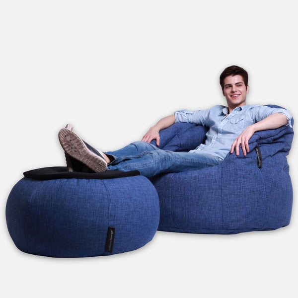 En ung man i jeansskjorta och jeans kopplar av på en Ambient Lounge Cove Package, en designad sittgrupp med bekväma möbler, med fötterna upplagda på en matchande blå ottoman och ler mot kameran mot en vit bakgrund.