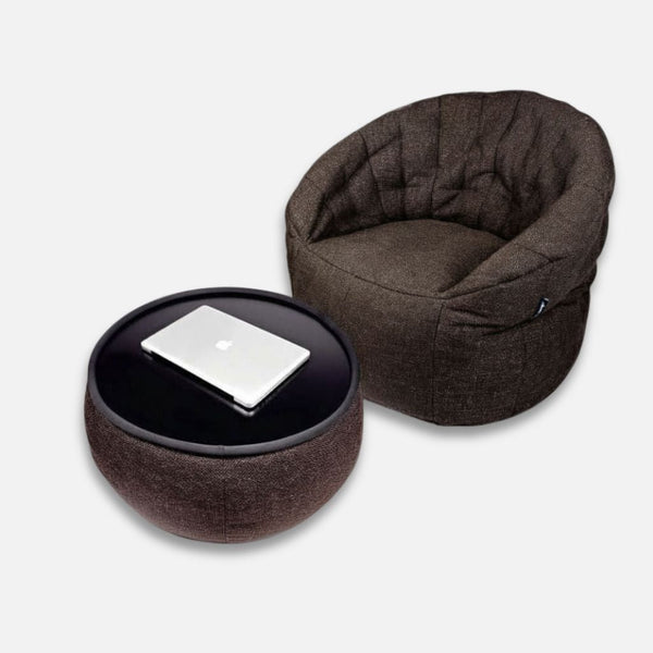 En brun, rund, stoppad stol från Ambient Lounge Cove Package står bredvid en matchande ottoman, som används som bord med en stängd silverfärgad laptop på toppen. Båda möblerna har ultramykt stoff för bekväma möbler mot en vit bakgrund.