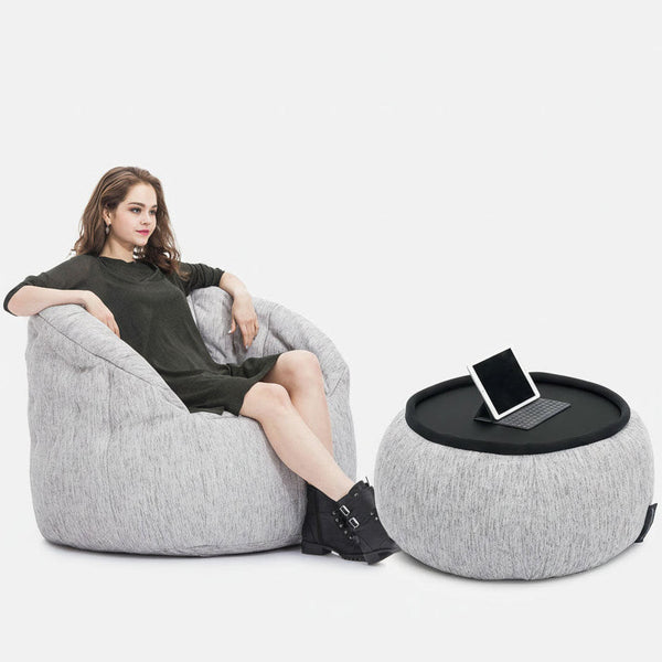 En kvinna i mörk klänning slappnar av i en ljusgrå saccosäcksfåtölj från Ambient Lounge, med benen på en matchande ottoman med en svart bricka som håller en surfplatta. Den bekväma möbeluppsättningen står mot en vit bakgrund.