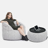 En kvinna i mörk klänning slappnar av i en ljusgrå saccosäcksfåtölj från Ambient Lounge, med benen på en matchande ottoman med en svart bricka som håller en surfplatta. Den bekväma möbeluppsättningen står mot en vit bakgrund.