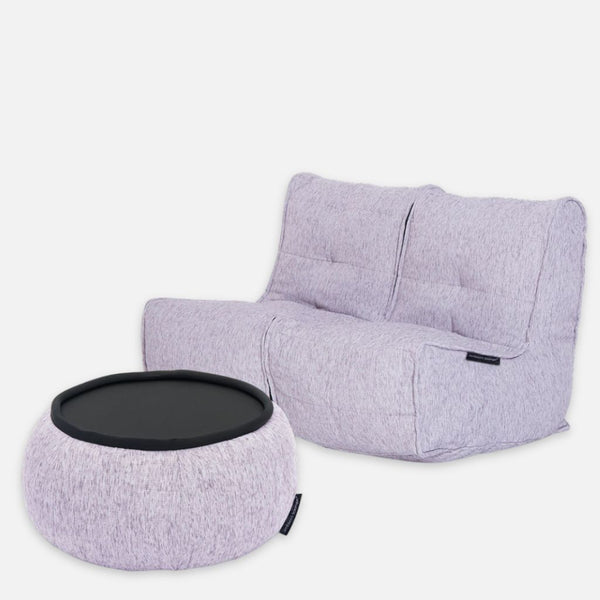 En modern, ljusgrå tvillingsoffa i tyg med tuftade kuddar och en matchande rund ottoman med svart brickskiva, båda mot en ljusgrå bakgrund.