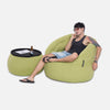 En man i svart linne, shorts, flip-flops och glasögon sitter på en grön saccosäckstol i Sunbrella-stoff - en del av en elegant utemøbler - med en matchande ottoman som bord, håller i en ölflaska och vilar huvudet på handen.