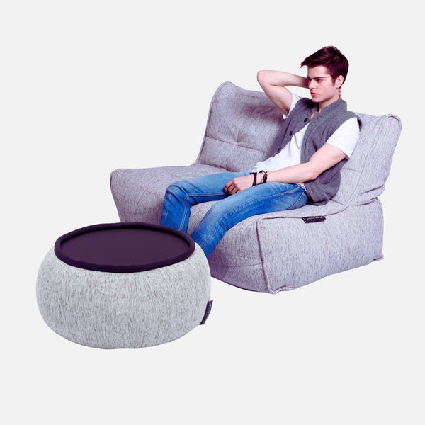 En ung man sitter avslappnat i en modern, ljusgrå flexibel soffa med en arm vilande på ryggstödet. Framför honom står en matchande rund ottoman med svart bordsskiva, båda på en vit bakgrund.