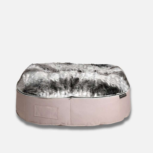 En rund hundeseng med en bas i ljusbeige tyg och en tjock, plyschig topp i fuskpäls i nyanser av grått, svart och vitt, perfekt för mysiga tupplurar - mot en vit bakgrund.