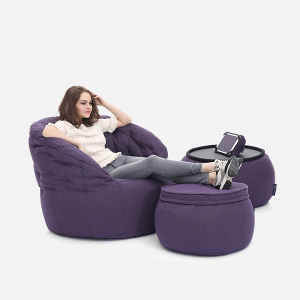 En kvinna slappnar av i en stor lila saccosäck med fötterna på en matchande ottoman - en del av Contempo Indoor Package - bredvid ett litet runt bord, som alla framhäver bekväma möbler och modern design mot en vit bakgrund.