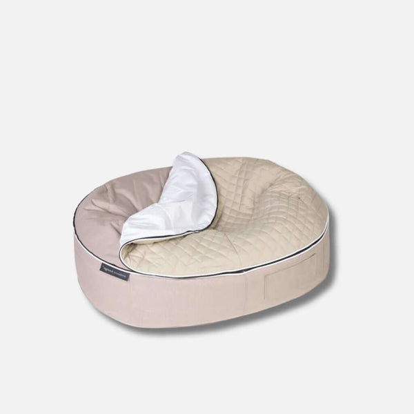 En beige, ovalformad allergivennlig hundeseng med quiltat topptrekk och en vit, delvis hopvikt filt på ena sidan, placerad på en ljusgrå bakgrund.