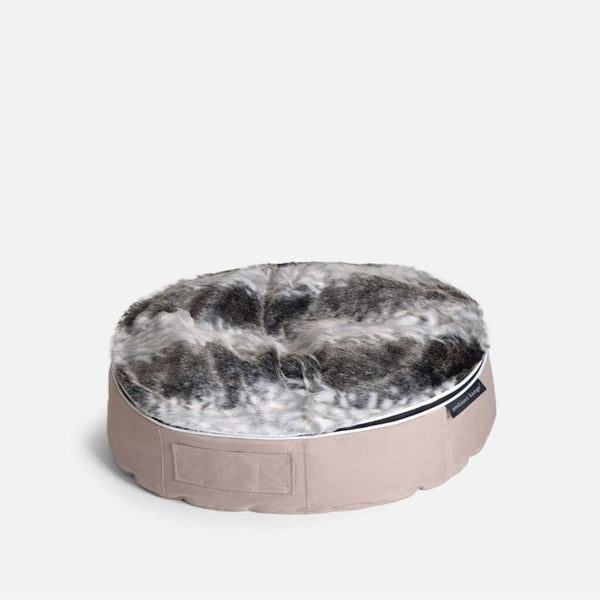 En rund hundeseng med en beige tygbotten och en plyschig topp i fuskpäls i nyanser av grått, vitt och svart erbjuder en elegant och bekväm hundeseng för ditt husdjur, vackert placerad på en vit bakgrund.