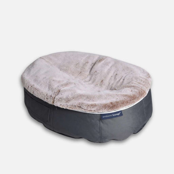 En mjuk, oval hundeseng med en mjuk, ljusbeige dyna och ett mörkgrått tygunderrede, med en liten Ambient Lounge-etikett på sidan för ultimat komfort för hund.