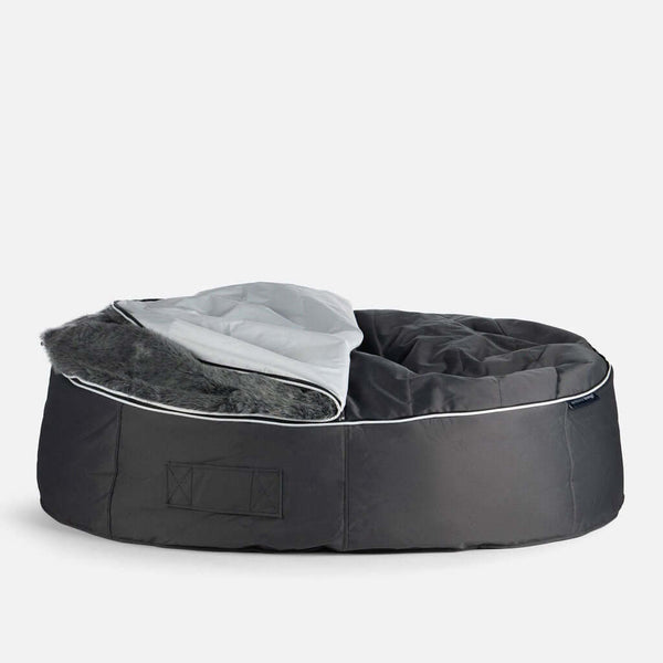 En stor, rund, mörkgrå bädd för husdjur med upphöjd kant och avtagbart överdrag av grå varg, med en del av det mjuka fodret fuskepelstrekk hundeseng delvis vikt bakåt, mot en vit bakgrund.