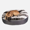 En stor brun hund ligger på en Ambient Lounge hundeseng XL, en rund, plyschig hundbädd med grå och vit fuskpäls på ovansidan och en mörkgrå tygbotten, mot en vit bakgrund.
