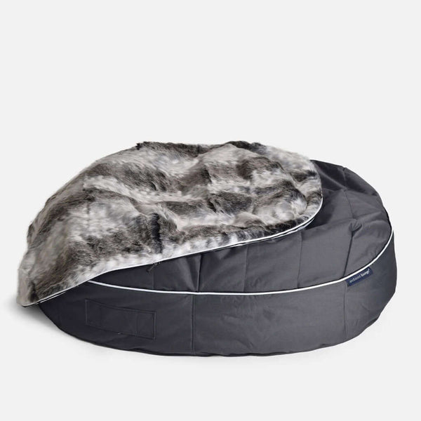 En stor, rund, mörkgrå hundeseng med en luksuriøs pelstopp fuskepelstrekk delvis draperad över sig, mot en enfärgad vit bakgrund.