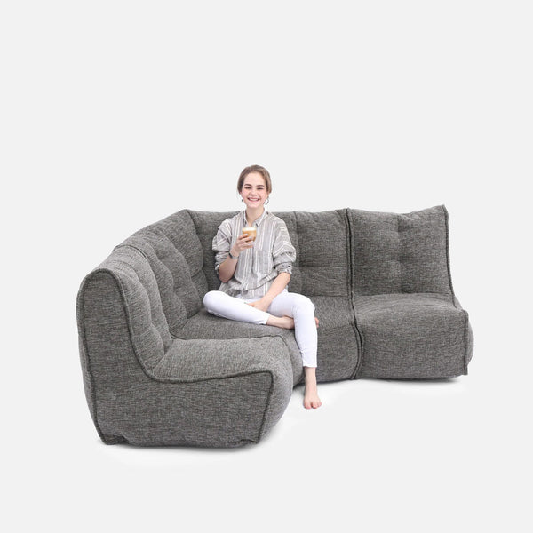 En leende kvinna sitter med benen i kors på en grå, modern soffa och håller en drink i handen mot en vit bakgrund.