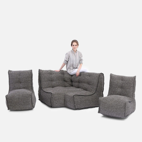 En kvinna i ljus randig skjorta sitter på ryggstödet till en modern, grå modulært hjørnemøbel med två matchande fåtöljer, alla med en mjuk, tuftad design mot en vit bakgrund.