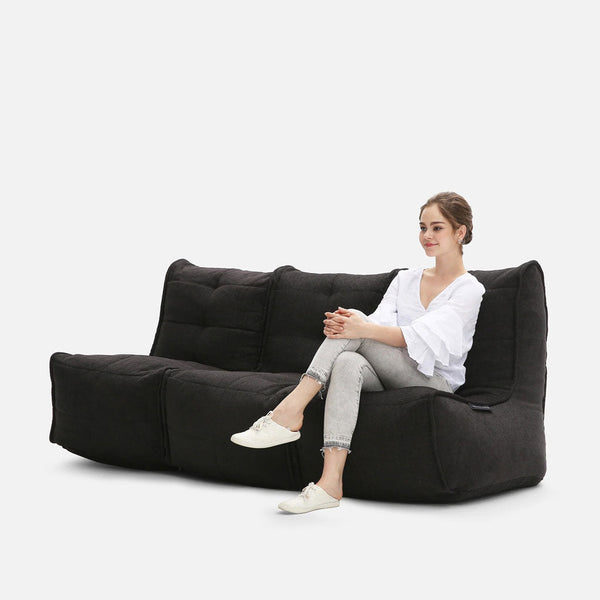 En kvinna i vit blus och grå byxor sitter med benen i kors på en modern myke møbler Mod 3 Movie Couch, vars svarta, tuftade design sticker ut mot den vita bakgrunden.