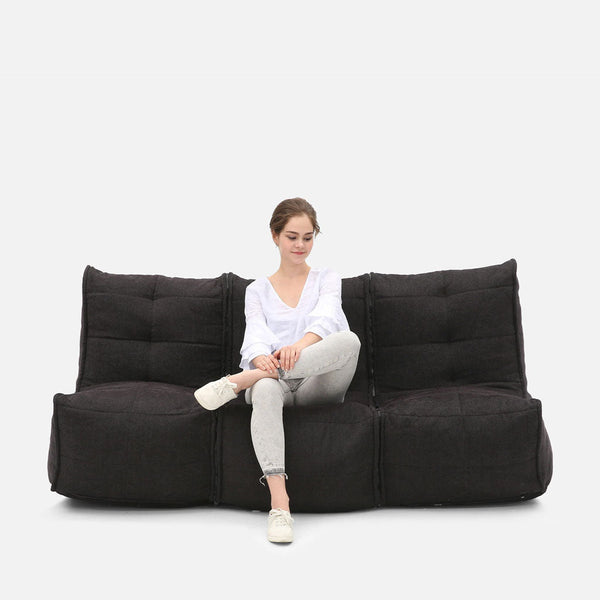 En kvinna i vit blus och ljusgrå jeans sitter med benen i kors på en modern, mörkgrå, vadderad Mod 3 Movie Couch från myke møbler mot en vit bakgrund.