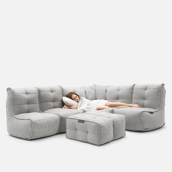 En kvinna i vit klänning ligger på en ljusgrå, tuftad Modulsofa med matchande ottoman och vilar huvudet på en kudde i en avslappnad position. Den mångsidiga designen kombinerar komfort och stil och är perfekt för moderna utrymmen. Bakgrunden är enfärgad vit.