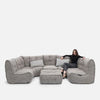 En kvinna sitter i hörnet av en stor, modern Ambient Lounge modulær-soffa med plyschkuddar och två matchande ottomaner, vilket skapar en elegant modern stueløsning mot en vit bakgrund.