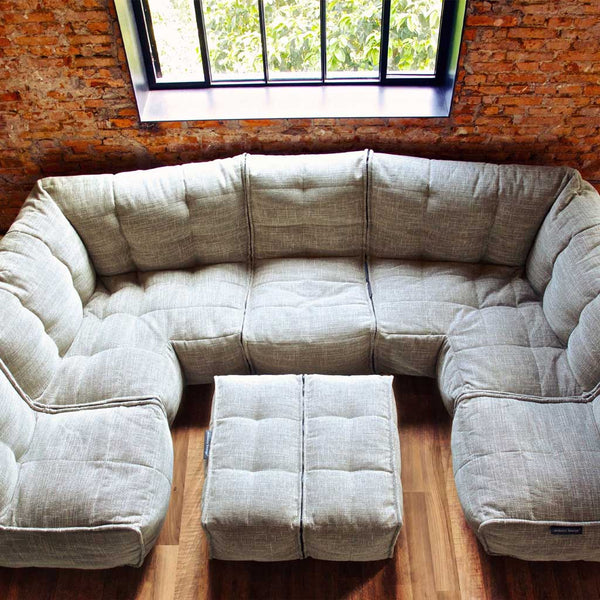 En ljusgrå, tuftad modulærsoffa från Ambient Lounge med en ottoman står på ett trägolv nära ett stort fönster med svarta karmar och en tegelvägg, med utsikt över gröna träd utanför.