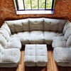 En ljusgrå, tuftad modulærsoffa från Ambient Lounge med en ottoman står på ett trägolv nära ett stort fönster med svarta karmar och en tegelvägg, med utsikt över gröna träd utanför.