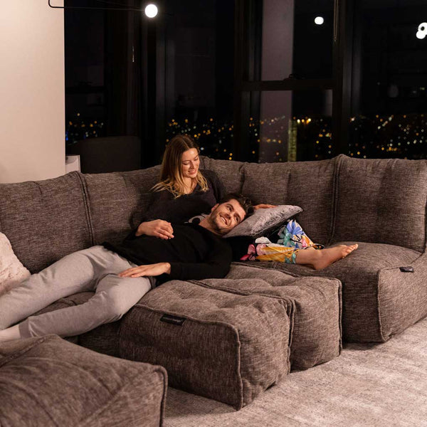 Ett par kopplar av i en stor grå Ambient Lounge modulær-soffa på kvällen. Mannen ligger med huvudet i kvinnans knä och ler när hon försiktigt vilar sin hand på hans bröst. Stadens ljus skiner in genom fönstren i denna moderna stueløsning.