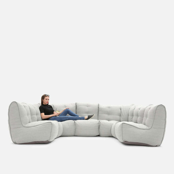 En person sitter avslappnat med benen utsträckta på en stor, modern Ambient Lounge sektionssoffa med tuftade kuddar mot en vit bakgrund.