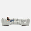 En person sitter avslappnat med benen utsträckta på en stor, modern Ambient Lounge sektionssoffa med tuftade kuddar mot en vit bakgrund.