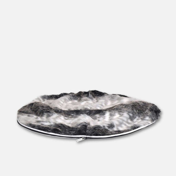 En rund, platt hundeseng med en fluffig, svart, vit och grå fuskepelstrekk och luksuriøs pelstopp, mot en enfärgad ljusgrå bakgrund.