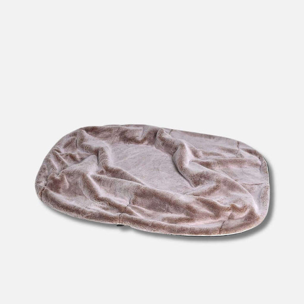 En mjuk, oval hundeseng med ljusbrun fuskepelstrekk och lätt skrynklig yta, mot en enfärgad vit bakgrund.