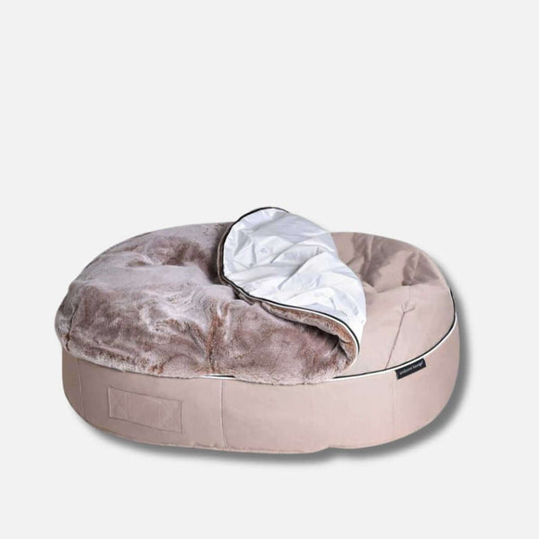 En rund, beige hundeseng med en plyschig, ljusbrun fuskepelstrekk-yta och en delvis vänd vit, fodrad filt ovanpå. Sängen har en mjuk, vadderad botten och står på en slät, ljus bakgrund.