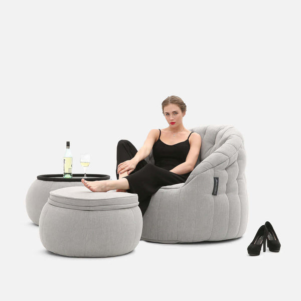 En kvinna i svart klädsel slappnar av barfota i en ljusgrå fåtölj med matchande ottoman och sidobord från Contempo Indoor Package, med en flaska vin och ett glas i handen. Svarta högklackade skor vilar på golvet nära ensemblen från Moderne Møbler.