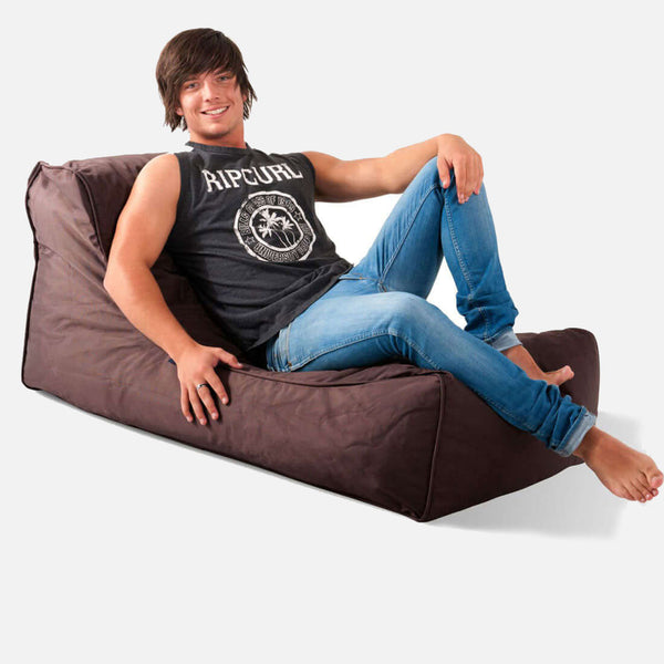 En ung man med brunt hår, klädd i en svart ärmlös Rip Curl-tröja och blå jeans, ler och lutar sig tillbaka på en brun Studio Lounger-säck mot en vit bakgrund.