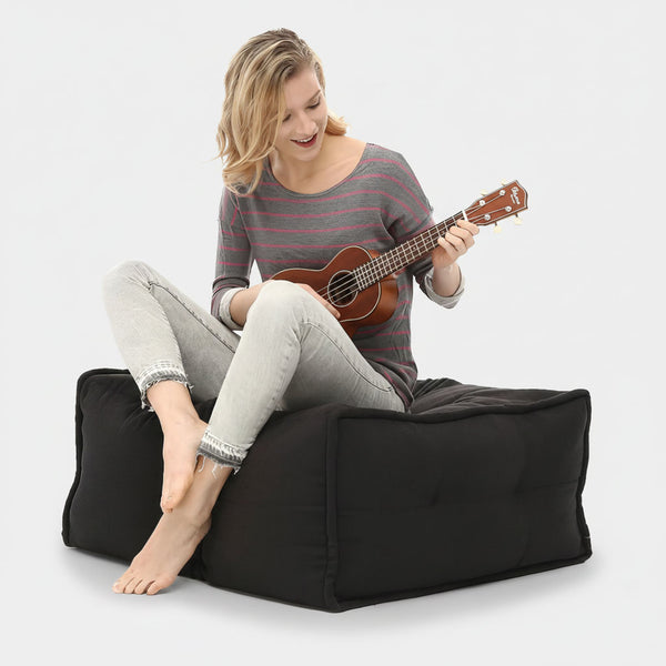 En person med blont hår, klädd i grårandig skjorta och ljusa jeans, sitter barfota på en Ambient Lounge Twin Ottoman och ler medan han spelar på ett litet stränginstrument, möjligen en ukulele - modern möblering när den är som bäst.