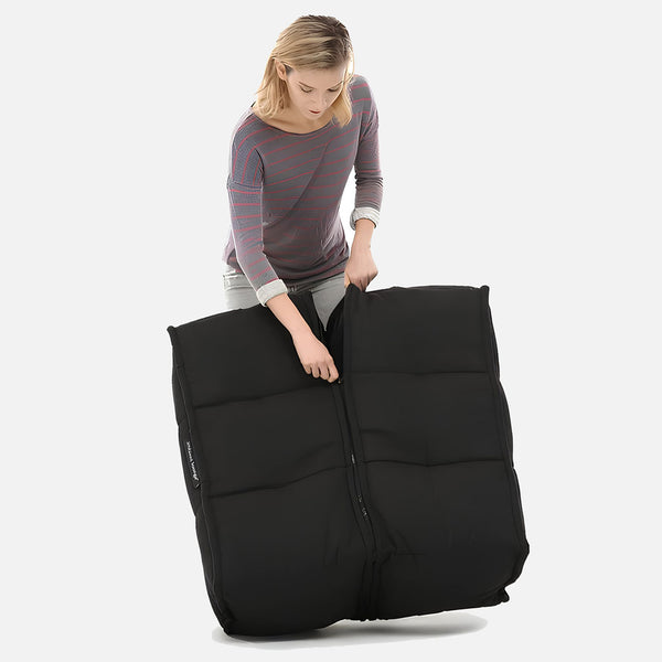 En person i randig långärmad skjorta och ljusa byxor lyfter en stor, svart, vadderad Twin Ottoman med dragkedja - från Ambient Lounge - mot en vit bakgrund och visar upp en stilren modern möbel.