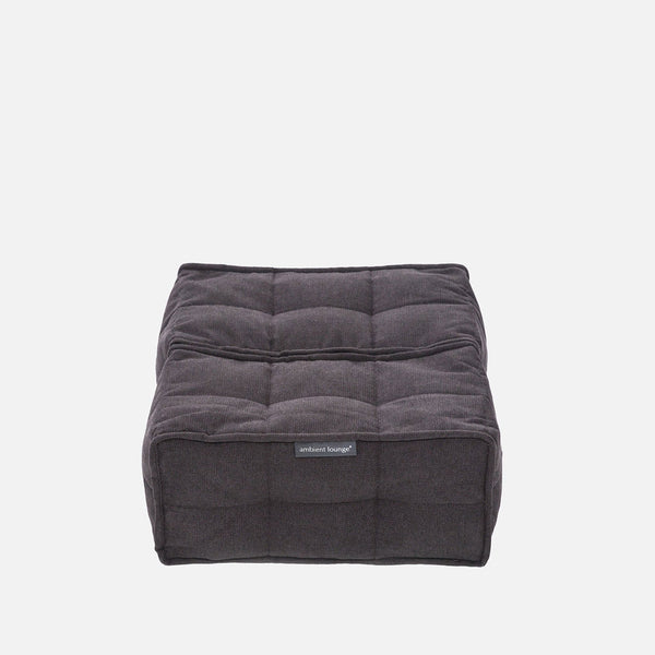 En fyrkantig, mörkgrå, tuftad Twin Ottoman från Ambient Lounge med en mjuk tygstruktur och en liten etikett på framsidan, som visar modern möbelstil mot en vit bakgrund.